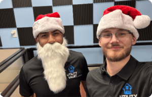 Two people wearing Santa hats, one with a fake white beard, in a room with checkered acoustic panels. Both are in black shirts with the "Utility Valet" logo.