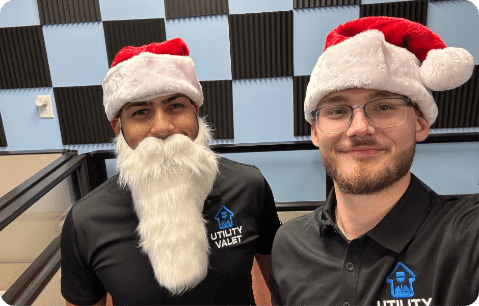 Two people wearing Santa hats, one with a fake white beard, in a room with checkered acoustic panels. Both are in black shirts with the "Utility Valet" logo.