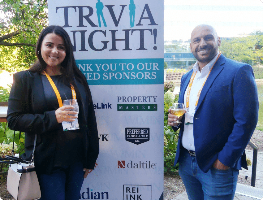 Two people smiling and holding drinks in front of a "Trivia Night" sponsor banner outdoors.