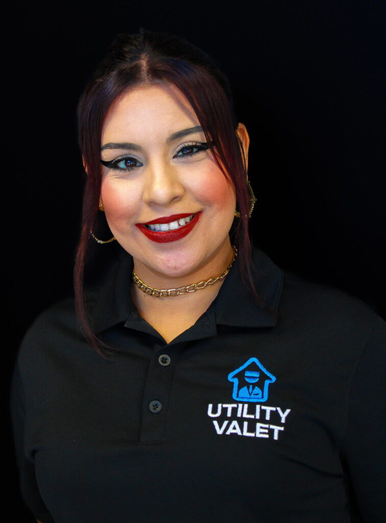 Woman wearing a black Utility Valet polo shirt, gold hoop earrings, and a gold chain necklace, smiling in front of a black background.