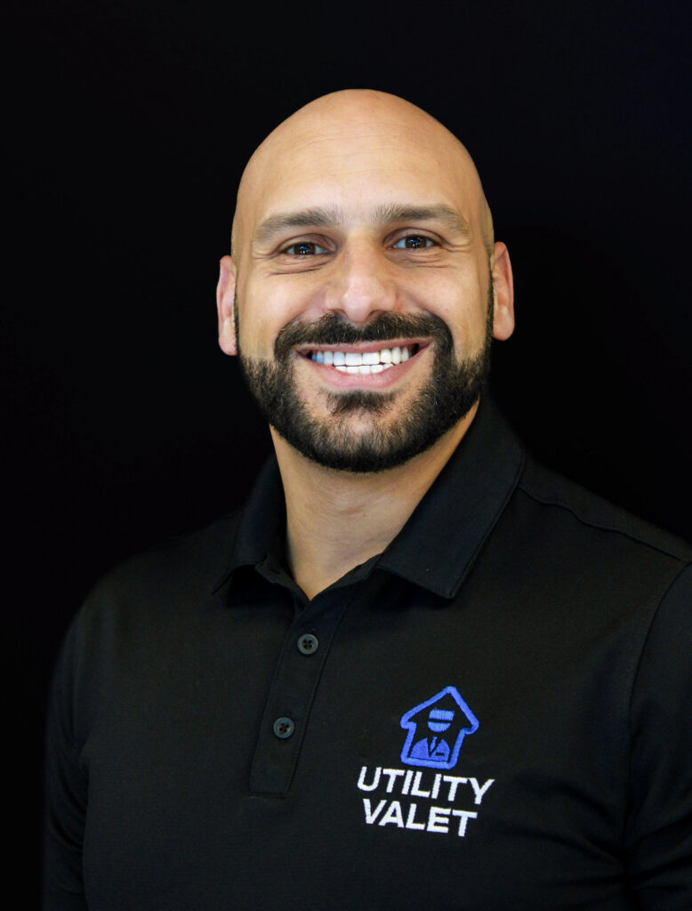 A man with a shaved head and beard wearing a black Utility Valet polo shirt smiles in front of a plain black background.