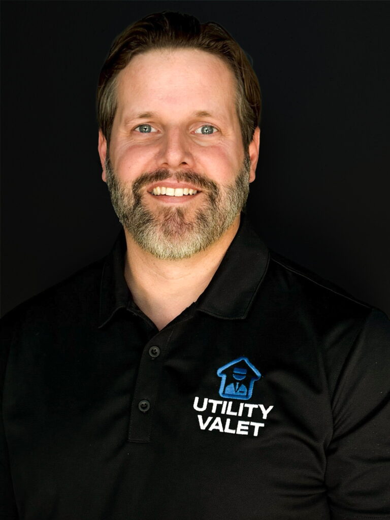 A man with brown hair and a beard smiles at the camera, wearing a black "Utility Valet" polo shirt, against a plain black background.