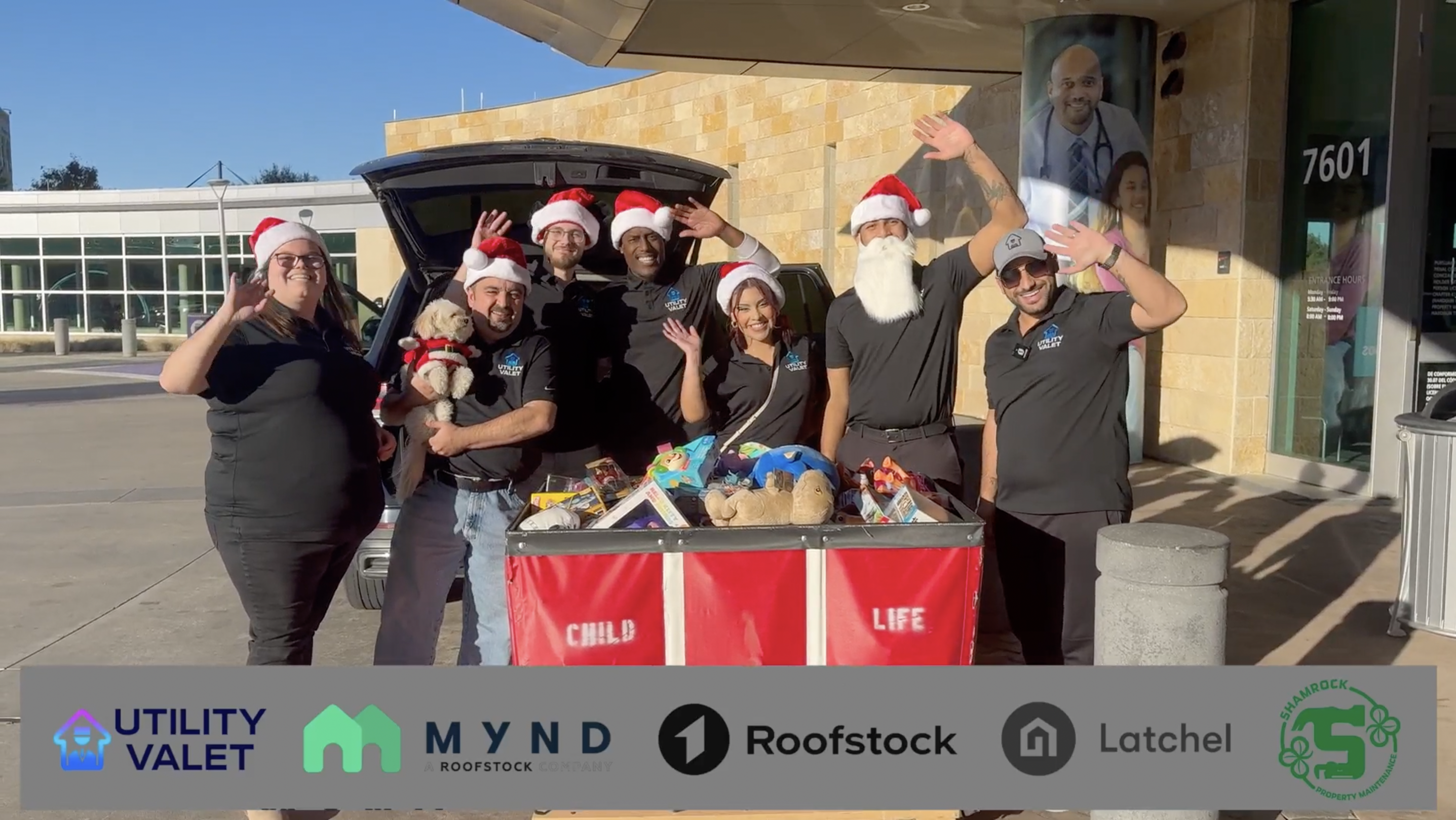 A group of people in black shirts and Santa hats stand outside by a cart filled with toys, smiling and waving. Sponsor logos appear at the bottom, highlighting community spirit and About Us - Utility Valet’s commitment to giving back.
